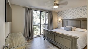 A bedroom with a large window, a bed with a wooden headboard, a ceiling fan, and a nightstand. - Stunning Pool Views! Close to Coligny Plaza/Beach (Hilton Head Island)