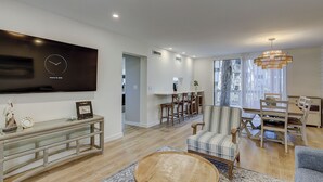 A modern living room with a flat-screen TV, wooden coffee table, striped armchair, and a dining area with bar stools. - Stunning Pool Views! Close to Coligny Plaza/Beach (Hilton Head Island)
