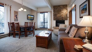 Un salón acogedor con chimenea de piedra, una zona de comedor con sillas de madera y un ventanal con cortinas.