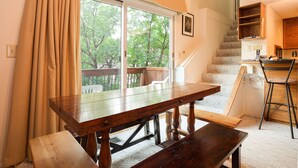 A room with a wooden dining table, a bench, a staircase, and a view of trees through a large window.