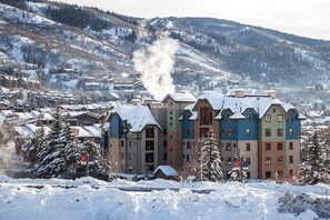 A snow-covered building with a chimney emitting smoke, surrounded by snow-laden trees and a mountainous backdrop.
