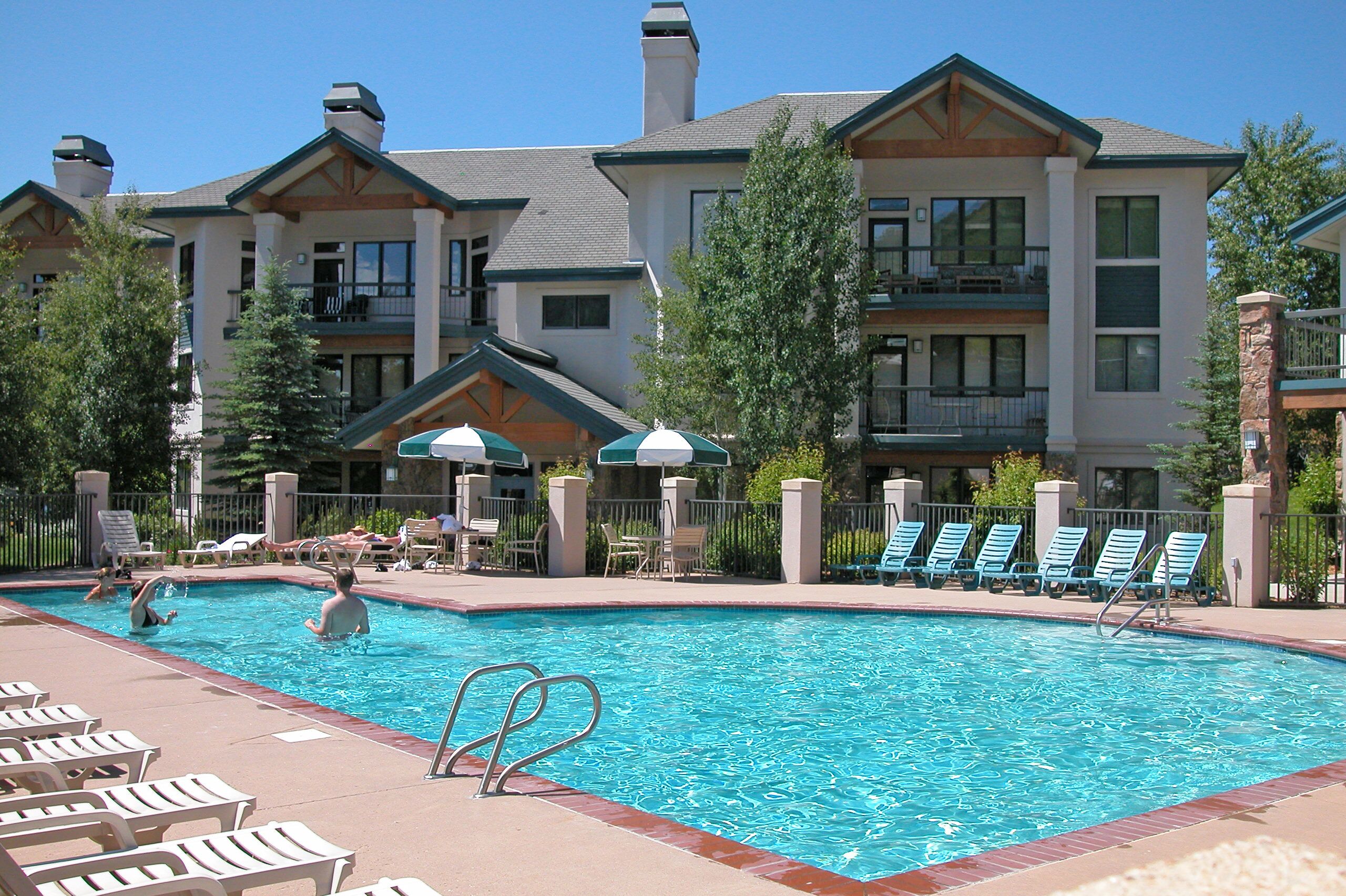 A swimming pool with lounge chairs and umbrellas, surrounded by a building with multiple balconies.