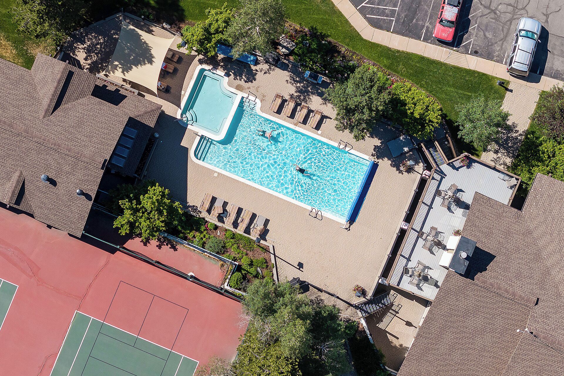 Aerial view of the property featuring a sparkling outdoor swimming pool surrounded by lounge chairs, tennis court, and landscaped green spaces with ample parking nearby.
