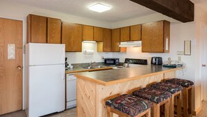 Cocina compacta con armarios de madera, un refrigerador blanco, microondas, tostadora y un lavamanos. Hay tres taburetes de madera con cojines estampados en la encimera.