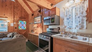 A cozy cabin-style kitchen with wooden walls, a stove, microwave, and a sink. - Moonridge Hideaway - Fireplace, Deck, Fenced Yard, Pet Friendly (Big Bear Lake)