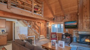 A cozy wooden cabin interior with a dining area, a living room with a fireplace, and a television mounted on the wall. - Moonridge Hideaway - Fireplace, Deck, Fenced Yard, Pet Friendly (Big Bear Lake)