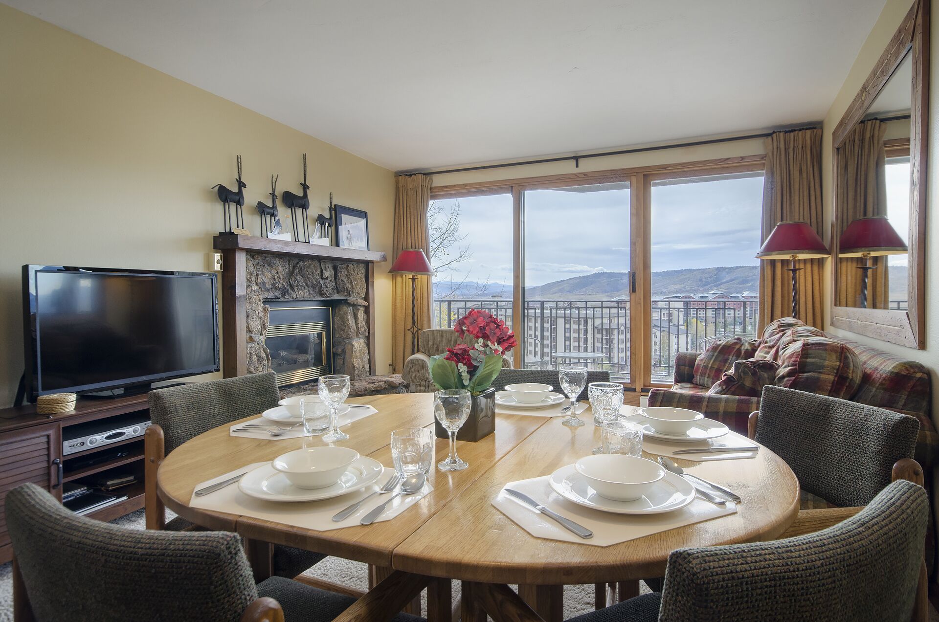 Un coin repas avec une table ronde dressée pour quatre personnes, une cheminée, un téléviseur et une vue sur les montagnes grâce à de grandes fenêtres.