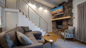 A modern living room with a brown leather sofa, a wooden coffee table, and a stone fireplace. A staircase with wooden railings leads to an upper level.