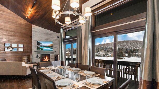 A dining area with a large wooden table set for six, a fireplace, and a view of a snowy landscape through large windows. - Managed by Park City Lodging-Premier Townhome in Lower Deer Valley-Ski Shuttle (Park City)