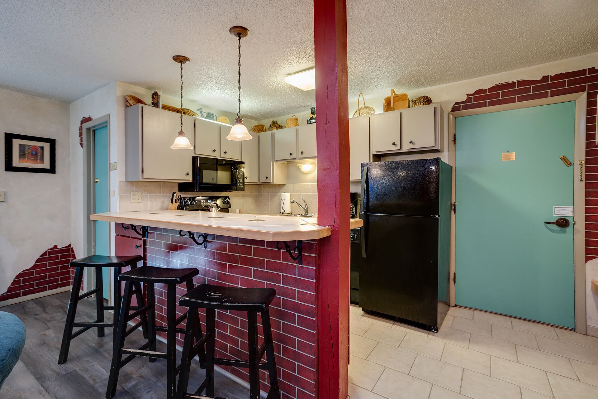 A compact kitchen with a black refrigerator, a microwave, and a sink. There are two stools and a small bar counter. The kitchen has a red brick accent wall and a teal door.