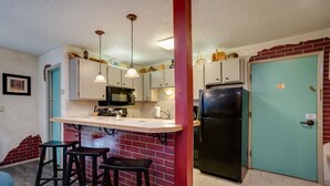 Cocina compacta con refrigerador negro, microondas y pileta. Hay dos taburetes y una pequeña barra. La cocina tiene una pared de ladrillo rojo y una puerta verde azulada.
