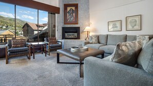A living room with a fireplace, a sofa, a chair, and a coffee table. There are framed pictures on the wall and a window with a view of buildings. - GSQ32 | Nice Views | Huge Private Deck | Steps from Steamboat Square (Steamboat Springs)
