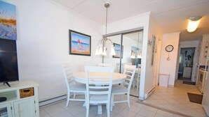 Un comedor con una mesa redonda y sillas, un televisor sobre un mueble bajo, y una cocina visible a través de una puerta abierta.