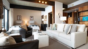 A hotel room with a large window, a white sofa, a black leather chair, and a white armchair. There is a wooden door and a wooden wall with a blue screen.