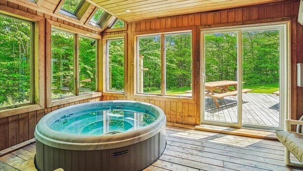 A hot tub in a wooden deck area with large windows and a view of a forest. - Sunburst home sleeps 16 with an indoor and outdoor hot tub.  Perfect for family reunions and getaways. (Killington)