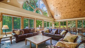 A spacious living area with leather sofas, a wooden coffee table, and large windows offering views of a forest. - Sunburst home sleeps 16 with an indoor and outdoor hot tub.  Perfect for family reunions and getaways. (Killington)