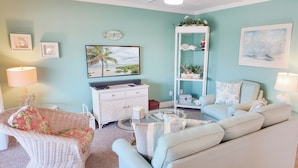 Una sala de estar con pared azul claro, un sofá blanco, una silla de mimbre, un televisor de pantalla plana, un armario blanco y una mesa de centro de vidrio.