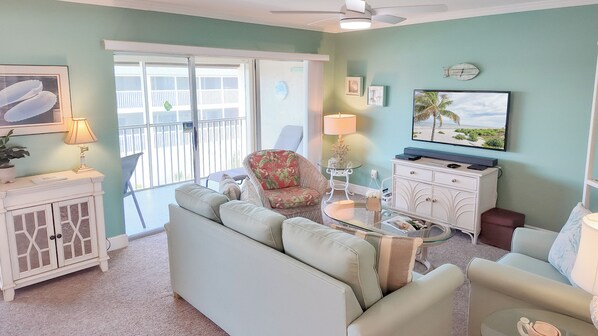 A living room with a sofa, armchair, and a small table with a lamp. There is a TV mounted on the wall and a window with a view of a balcony.