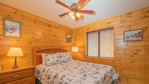 A wooden cabin room with a bed, a nightstand, a ceiling fan, and a window with blinds. - Moonridge Mountain - Hot Tub, Fenced Yard, BBQ, Walk to shops (Big Bear Lake)