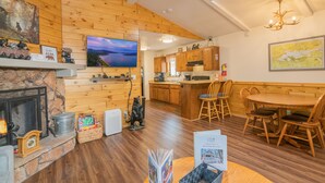 A cozy cabin interior with a fireplace, wooden walls, a dining area, and a kitchen. - Moonridge Mountain - Hot Tub, Fenced Yard, BBQ, Walk to shops (Big Bear Lake)