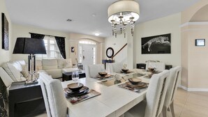 Une salle à manger avec une longue table dressée pour six personnes, entourée de chaises. Un lustre est suspendu au-dessus de la table, et un escalier est visible au fond.