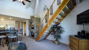 A spacious living area with a stone fireplace, a wooden staircase, a dining table with chairs, and a television on a wooden cabinet.