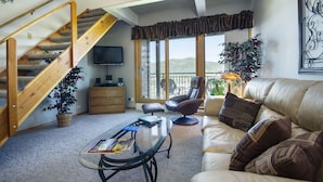 A spacious living room with a wooden staircase, a leather sofa, a glass coffee table, and a television mounted on the wall.