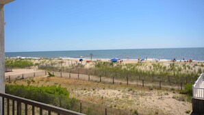 A beach view from a balcony with a wooden fence, beach umbrellas, and people in the distance. - Festive, fun 2 bedroom oceanfront condo with free WiFi, unique and sunny decor, and a beautiful view of the ocean located in quiet midtown and just a few steps to the beach! (Ocean city)