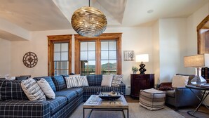 Un salon comprenant un canapé, une table basse et une lampe. Il y a des fenêtres avec des stores, une horloge au mur et une vue sur l’extérieur.