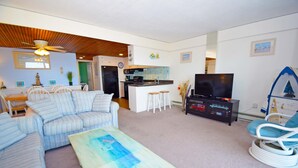 A living room with a sofa, a coffee table, a TV, and a kitchen area in the background. - Beachmark 3F-Oceanfront 73rd St, WIFI, Pool, Elev, W/D, AC (Ocean City)