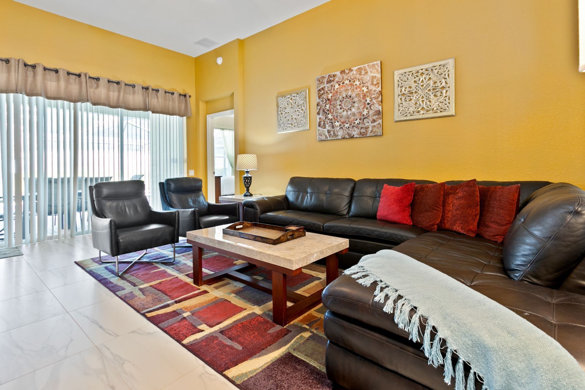 A living room with a black leather sofa, two armchairs, a coffee table, and a colorful rug. There are two wall-mounted artworks and a window with vertical blinds.