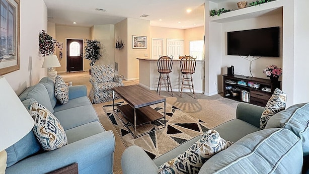 A spacious living room with a sofa set, a coffee table, and a TV. There is a dining area with chairs and a kitchen in the background.
