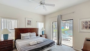 A bedroom with a large bed, a ceiling fan, a nightstand with a lamp, and a view of the backyard through a sliding glass door.