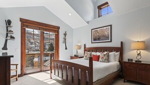 Une chambre à coucher avec un grand lit, une tête de lit en bois, une fenêtre donnant sur un paysage enneigé, un tableau au mur et une table de chevet avec une lampe.