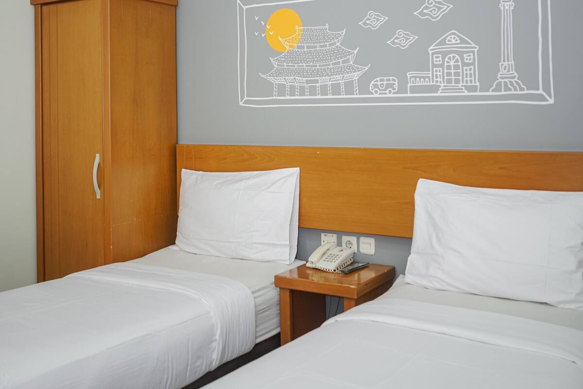 A hotel room with two beds, a wooden wardrobe, a wall with a cityscape mural, and a bedside table with a phone.