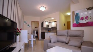 Un salon moderne avec un téléviseur à écran plat, un canapé beige, une table basse blanche et une cuisine en arrière-plan.