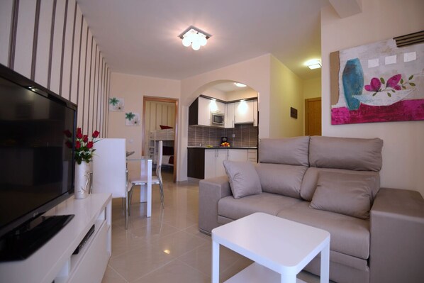 A modern living room with a flat-screen TV, a beige sofa, a white coffee table, and a kitchen in the background. - Apartment, WITH POOL VIEWS - NEXT TO THE BEACH-IDEAL FOR FAMILIES (Costa Calma)