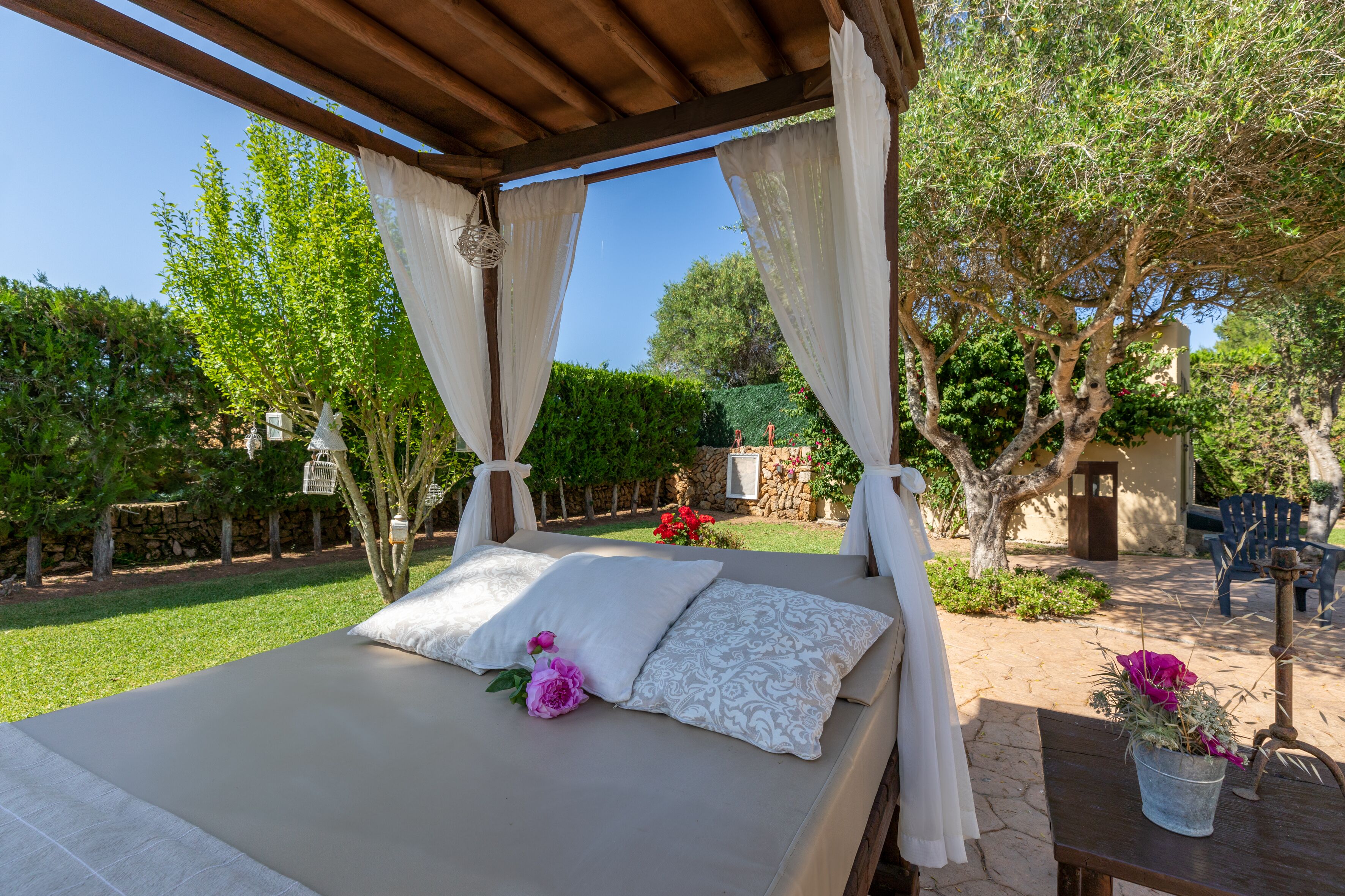 A bed with white and patterned pillows, a wooden table with a potted plant, and a pergola with white curtains.