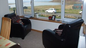 Una habitación con un gran ventanal con vistas a un lago y montañas, que incluye dos sillones de cuero negro, una computadora portátil y una mesita con un florero y un marco de fotos.