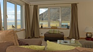 Una sala de estar acogedora con vista a un lago y montañas, amueblada con un sofá, reposapiés y una mesita con controles remotos.