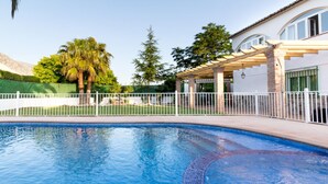 A swimming pool with a tiled edge, surrounded by a white fence, and a building with a pergola in the background. - Cottage with pool, garden with views and wifi (Dúrcal)