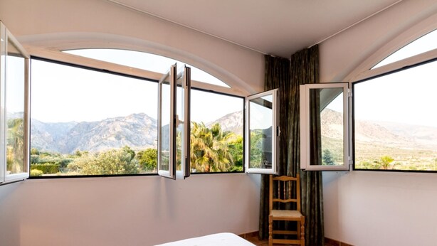 A bedroom with a large bed, two open windows, a wooden chair, and a view of mountains and greenery. - Cottage with pool, garden with views and wifi (Dúrcal)