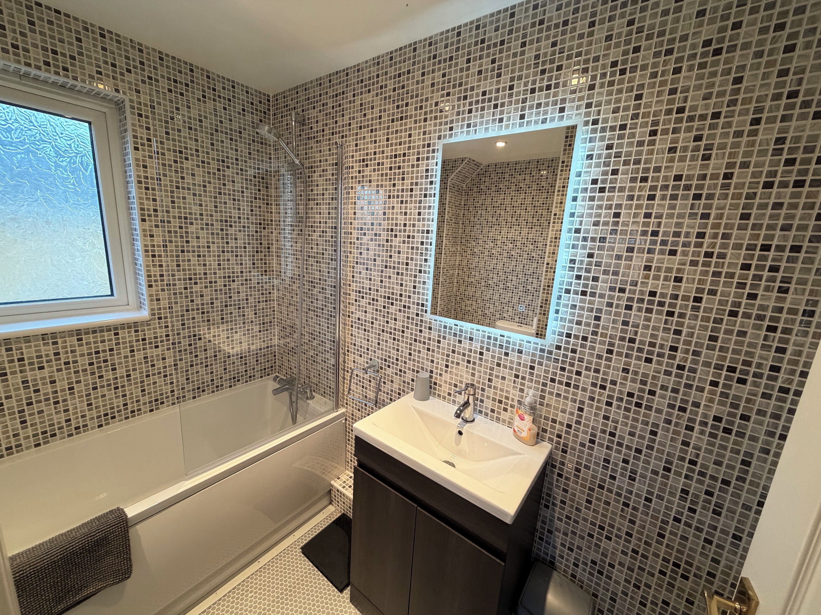 Une salle de bain moderne avec un mur recouvert d’une mosaïque, un grand miroir, une baignoire et un lavabo doté d’un distributeur de savon.