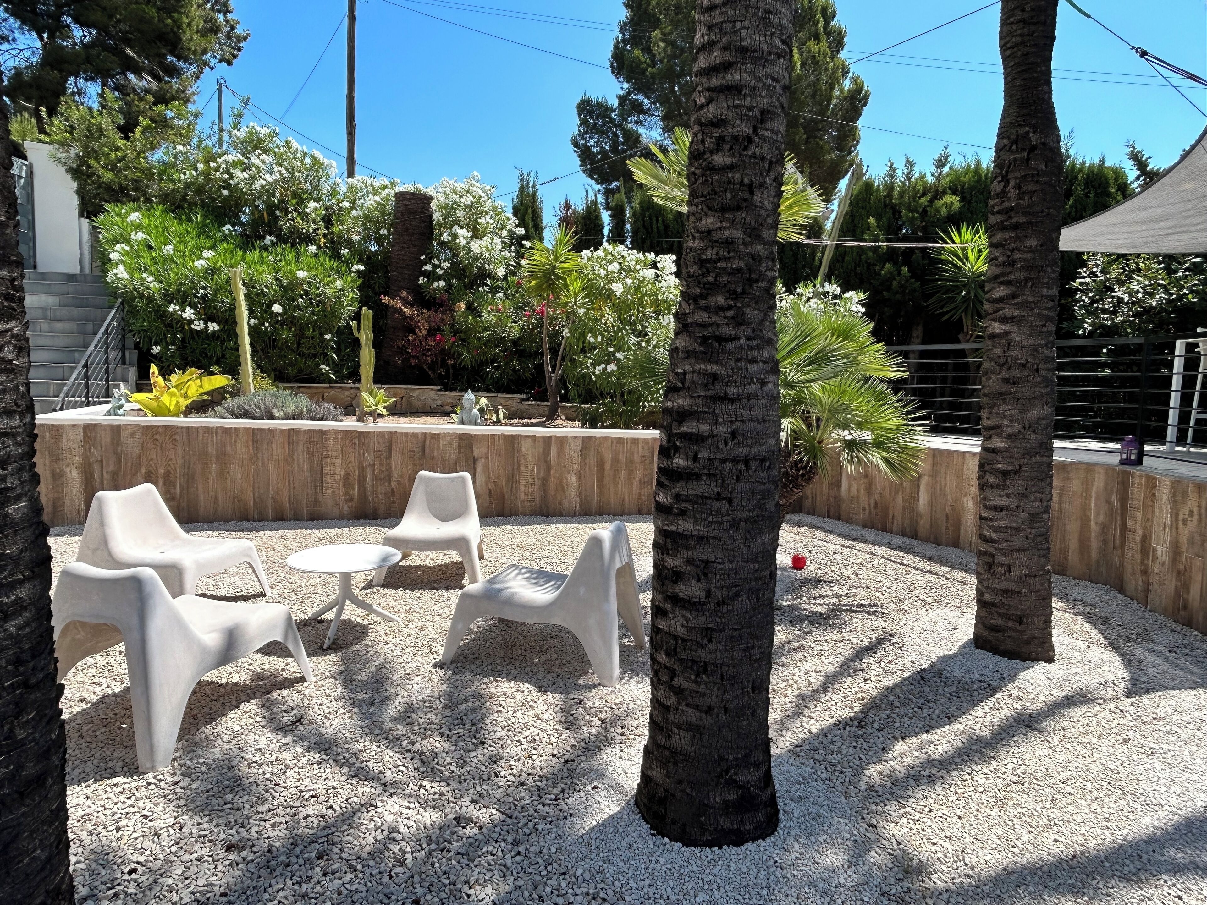 An outdoor seating area with white plastic chairs and a small round table, surrounded by gravel and palm trees.