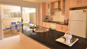 A modern kitchen with a black countertop, stainless steel sink, and a refrigerator. There is a dining area with a table and chairs visible through a glass door. - Adelaide Luxury Beach House (Henley Beach)