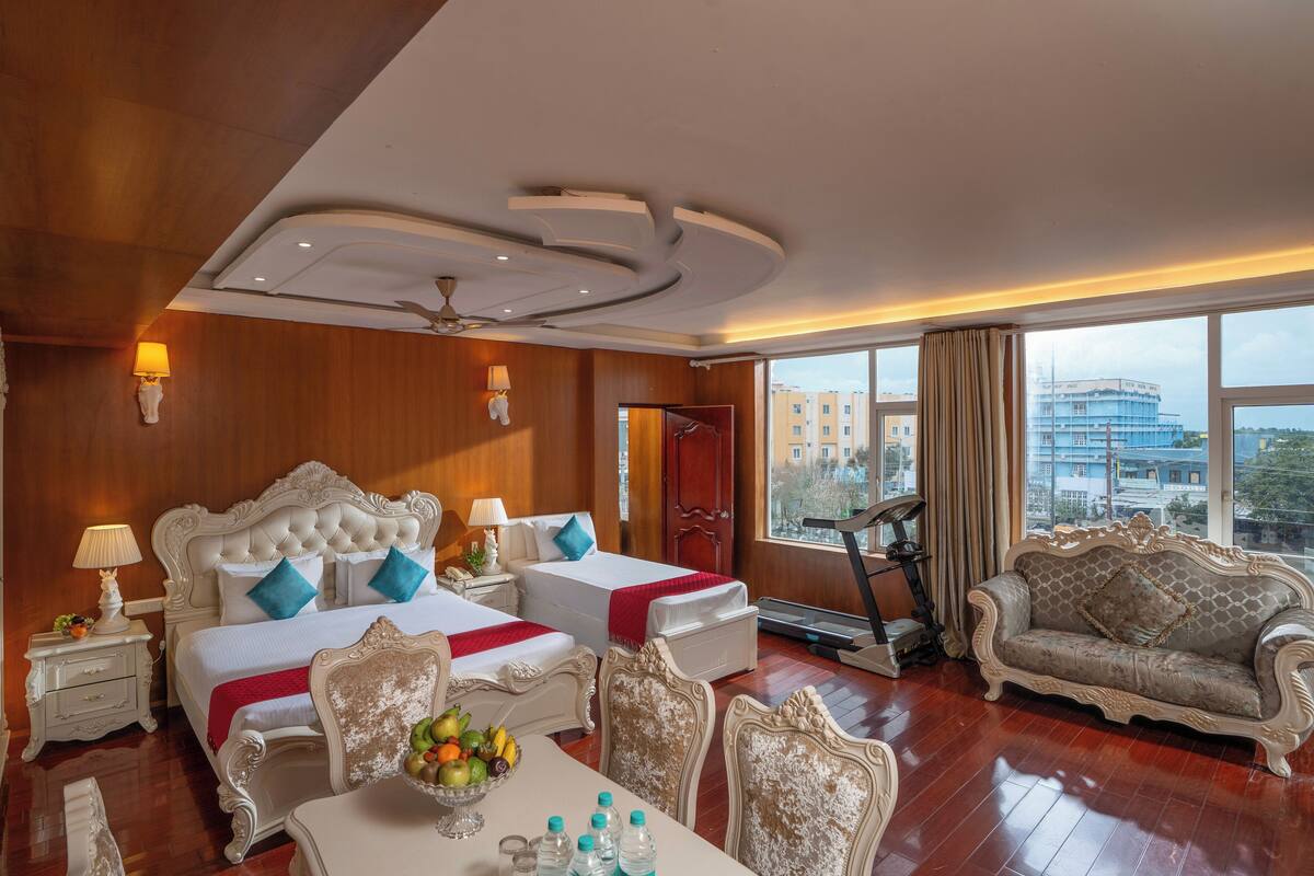 A hotel room with a large bed, two bedside tables, a dining table with fruit and water bottles, a sofa, and a view of the city through large windows.