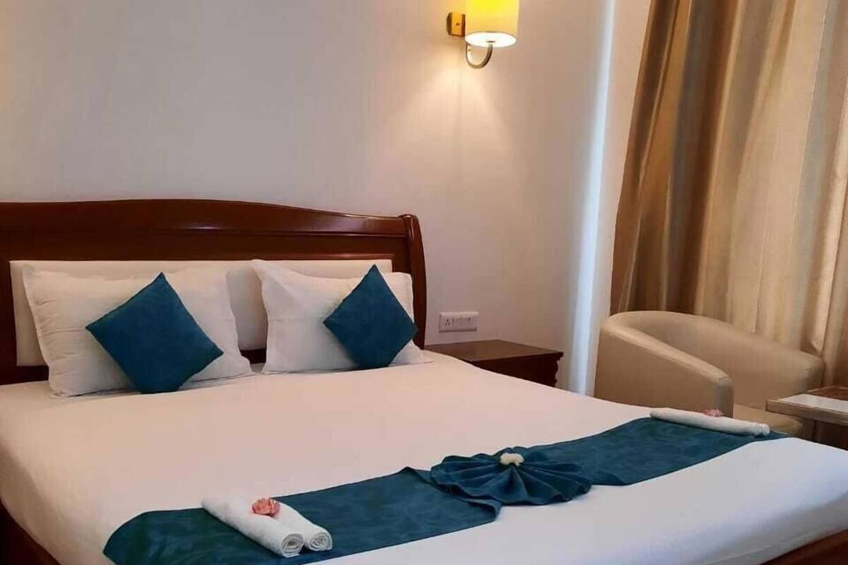A hotel room with a wooden bed, white bedding, a blue accent pillow, a wall-mounted light, a chair, and a curtain.