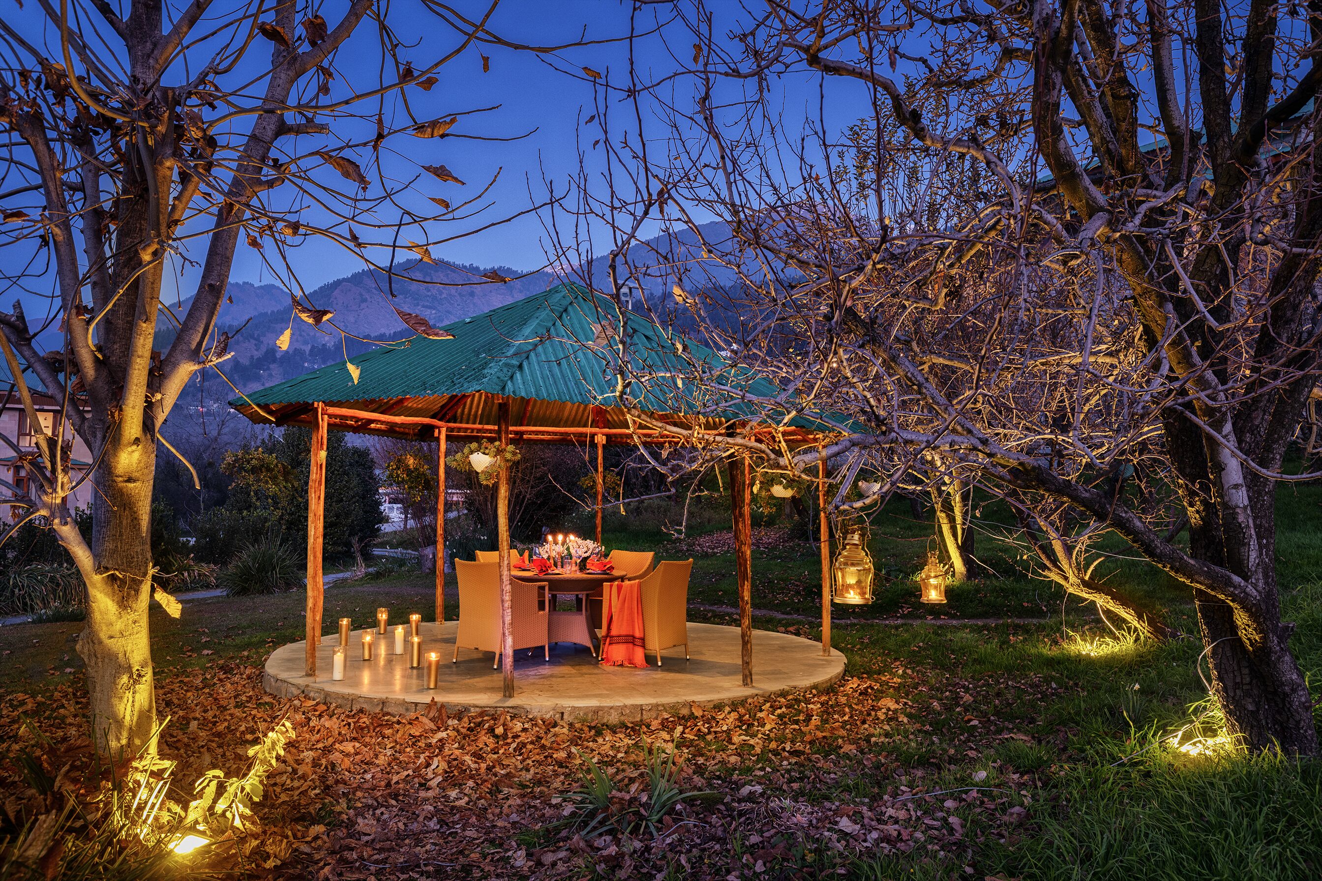 A circular outdoor dining area with a table and chairs, surrounded by trees and a mountain backdrop.