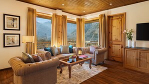 A cozy living room with a wooden ceiling, large windows with curtains, a sofa set, a coffee table with fruit, and a television on a wooden cabinet. - Baragarh Resort & Spa, Manali- IHCL SeleQtions (Kullu)
