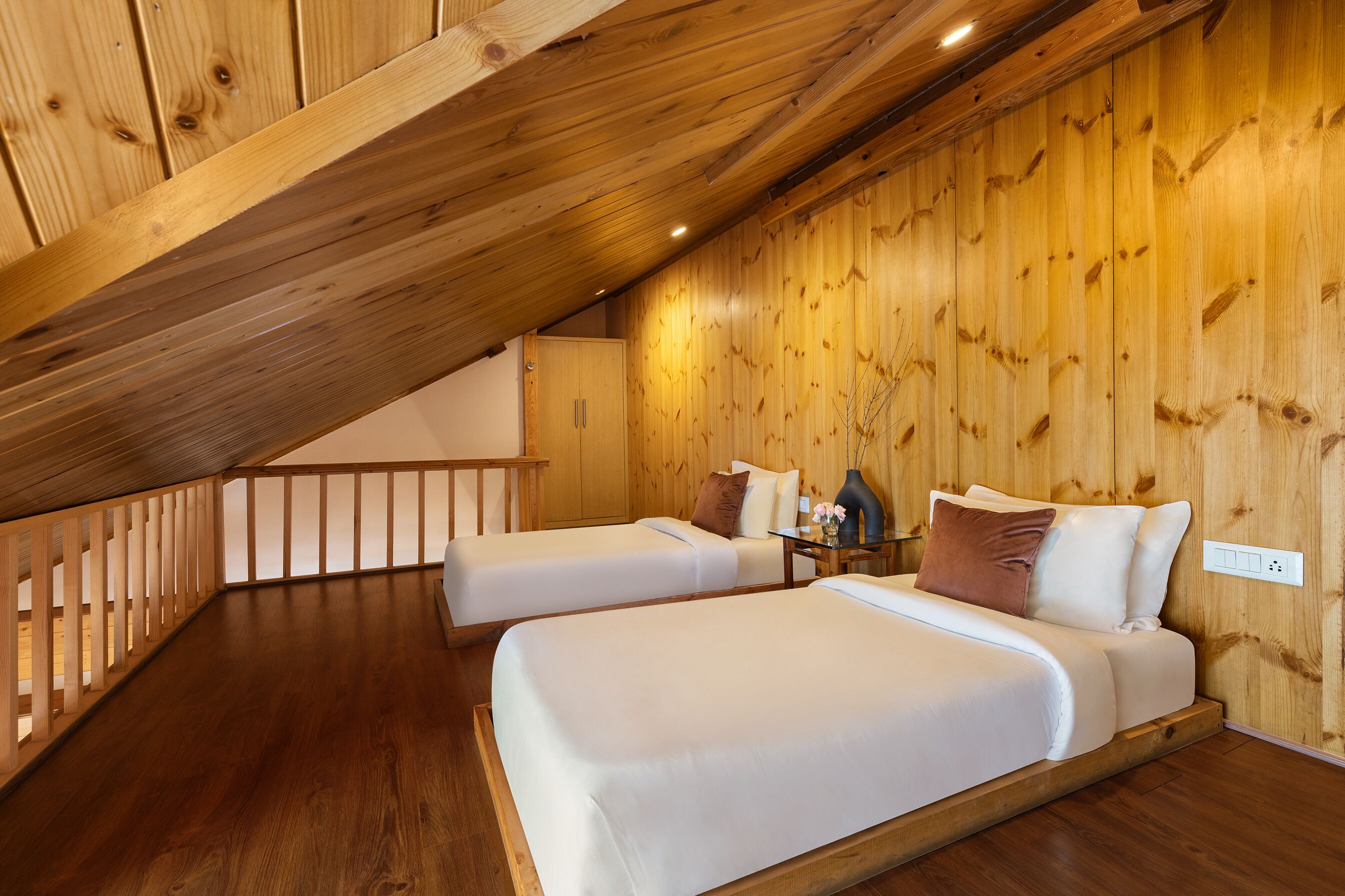 A room with two beds, wooden walls, a sloped ceiling, and a wooden railing.